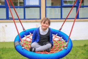 A young girl sits cross-legged on blue STEM playground equipment, a net swing, outdoors in front of a building with blue-framed windows.