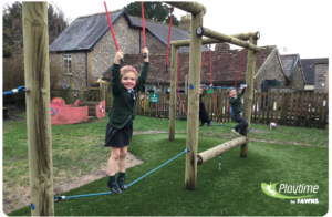 Three children play on a wooden balance and rope climbing structure in a grassy outdoor playground area, with buildings visible in the background.