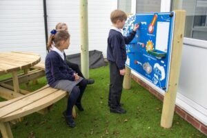 Three schoolchildren in uniforms use an outdoor science activity board, with two sitting at a round picnic table and one child standing and interacting with the board.