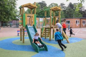 Children play on a playground structure with slides and climbing areas, surrounded by trees and a brick building in the background.
