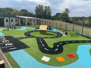 Outdoor play area with artificial grass, road markings, a small wooden house, climbing structures, and a fenced playground next to a building and open field.