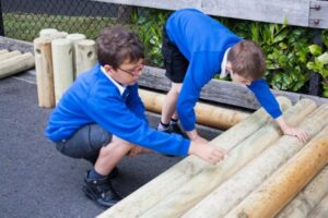 Two boys in school uniforms are arranging large wooden logs on the ground near a fence and some bushes.