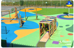Colourful outdoor playground with climbing frames, balance equipment, and a soft play surface, set in a fenced area next to a car park. Playtime by Fawns logo in the corner.