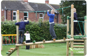 Four children in school uniforms play on a wooden playground structure featuring ropes and balance beams—perfect examples of outdoor fitness equipment for schools. Houses and trees are in the background. Logo in corner reads Playtime by FAWNS.