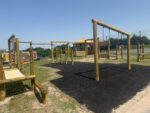 Outdoor playground with wooden climbing structures, swings, Inclusive Gym Rings, a rope bridge, and a gazebo. The ground has patches of grass and black safety mats under equipment. Fence and trees in the background.