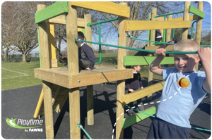Three children play on a wooden climbing frame outdoors under clear sky, with green grass and trees in the background. Playtime by FAWNS logo appears in the bottom left corner.