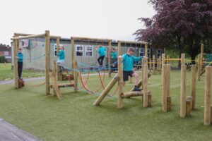 Children in school uniforms play on a wooden climbing frame with ropes in an outdoor playground area.