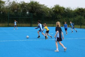 Six children play football on a blue outdoor court, with one child in yellow attempting to kick the ball while others look on or run nearby. Trees and a fence are in the background.