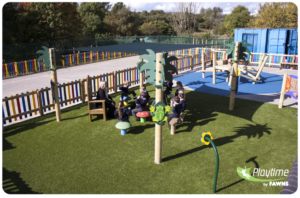 Several young children and an adult sit on wooden outdoor furniture in a fenced playground area with artificial grass and decorative play structures.