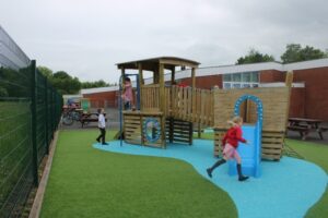 Three children play on a wooden playground structure with blue safety flooring, whilst another person sits at a nearby picnic table. The location is fenced and adjacent to a brick building.