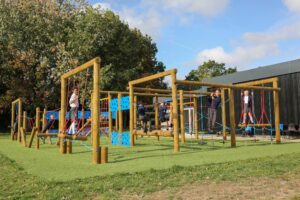 Children play on a wooden outdoor climbing structure with ropes and blue panels on a grassy area near a building, with trees in the background.