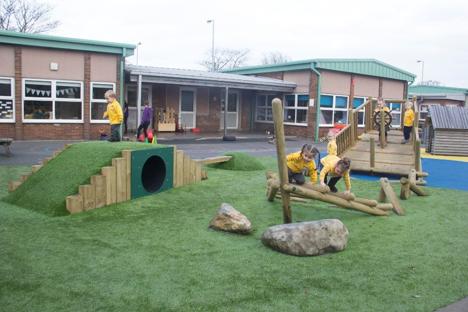 Children play on artificial grass with climbing equipment, rocks, and a tunnel outside a school building.