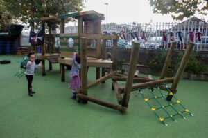 Three young children play on a wooden climbing frame in an outdoor playground with green rubber flooring and a fence in the background.