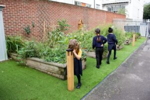 Three children in school uniforms stand and walk near raised garden beds along a brick wall in an outdoor area with artificial grass and a paved path.