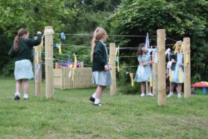 Several children in school uniforms navigate through strings tied between wooden posts in an outdoor play area, surrounded by grass and trees.