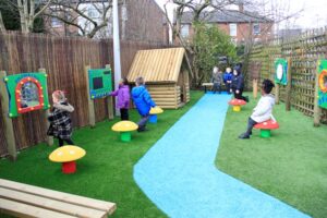 Children play and interact with activity boards and mushroom-shaped seats in a fenced outdoor play area with artificial grass and a blue path.
