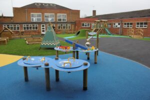 Outdoor school playground with blue activity tables, climbing frames, a slide, artificial grass, and brick buildings in the background.
