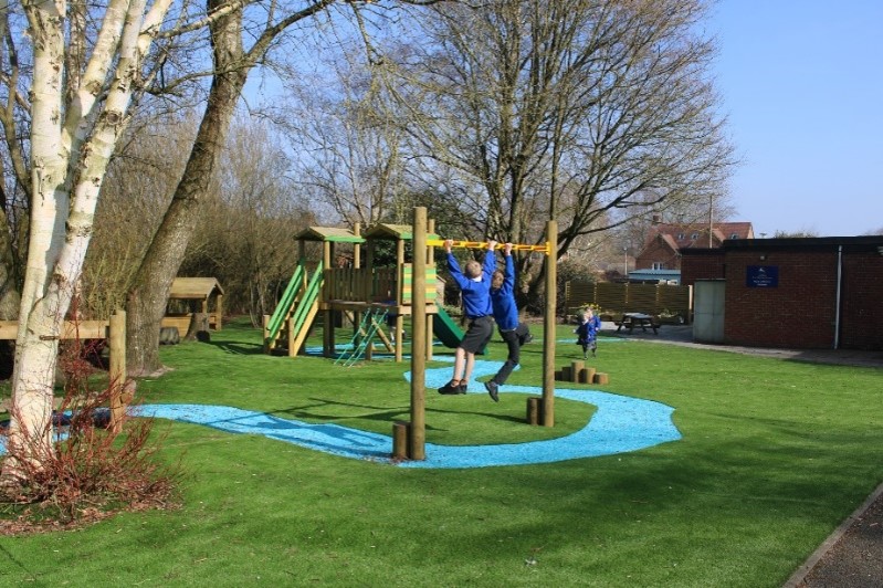 Two children use monkey bars on a playground with artificial grass, climbing structures, and trees on a sunny day, highlighting the importance of regular playground maintenance to ensure a safe and enjoyable environment.
