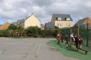 Children in school uniforms play on wooden playground equipment near a fenced area, with residential houses and cloudy skies in the background—a lively scene that highlights the value and investment behind school playground cost.