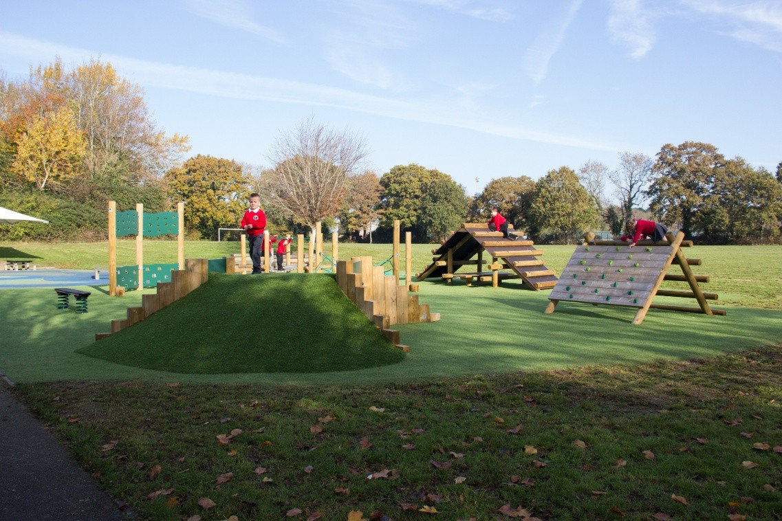 Several children in red uniforms play on a green outdoor playground with climbing frames and grassy areas, surrounded by trees and open field under a clear sky.