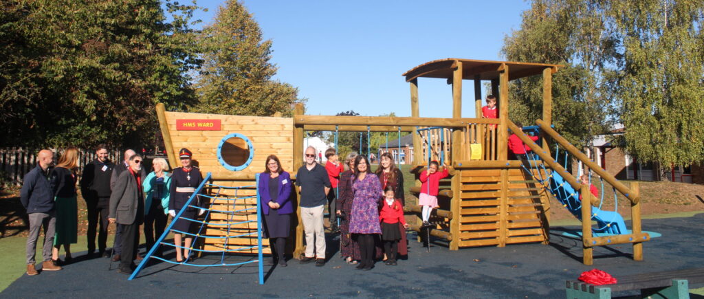 A group of adults and children from Hillborough Infant and Nursery School standing in front of a new wooden playground structure shaped like a boat, labelled HMS Ward.